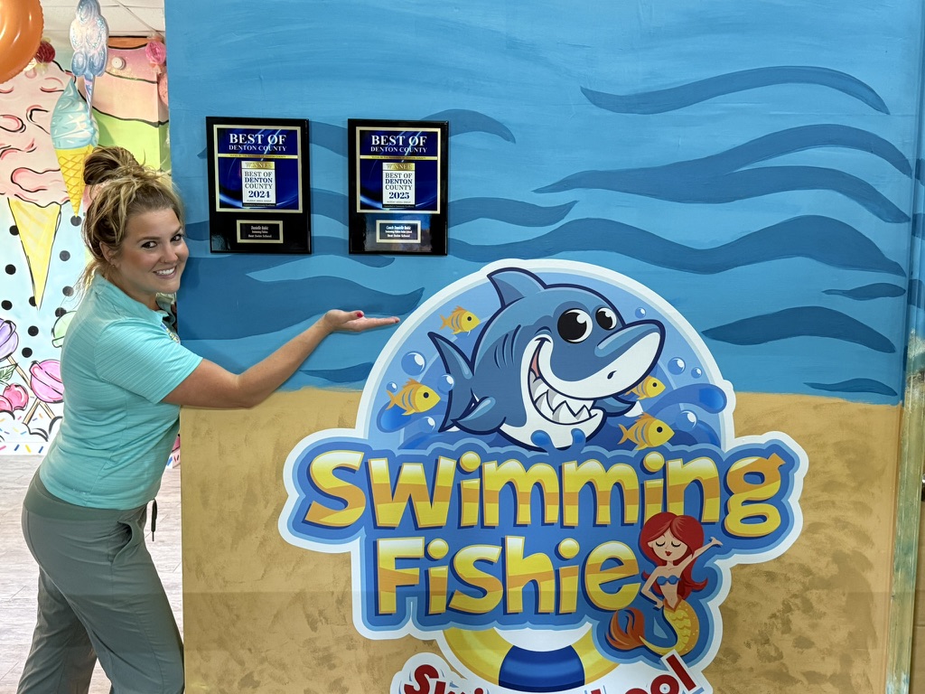 Coach posing beside Swimming Fishies awards and logo
